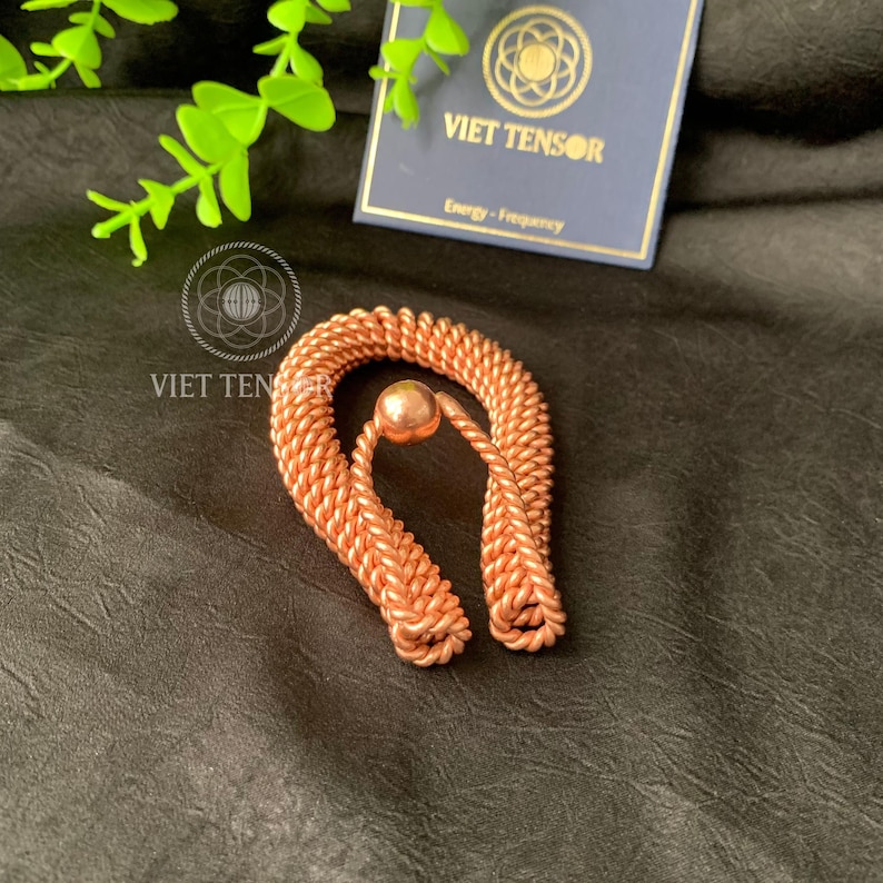 May include: A copper-colored, U-shaped metal object with a small sphere at the center. The metal is woven in a rope-like pattern. A business card with the text "VIET TENSOR" and "Energy - Frequency" is in the background.