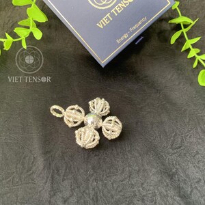 May include: A silver pendant with a four-petal design and a central bead. The pendant is on a black surface, with a blue box and green leaves in the background. The box has the text "VIET TENSOR" and "Energy - Frequency".