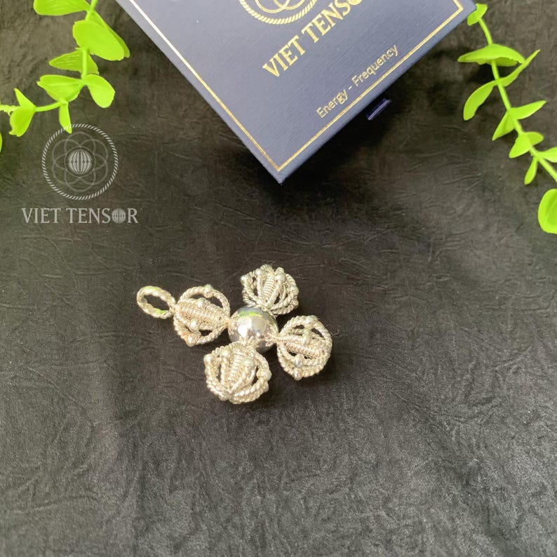 May include: A silver pendant with a four-petal design and a central bead. The pendant is on a black surface, with a blue box and green leaves in the background. The box has the text "VIET TENSOR" and "Energy - Frequency".