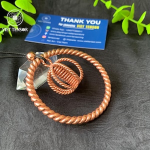 May include: A copper-colored pendant with a spiral design, featuring a large outer ring and a smaller inner ring with a cylindrical center. The pendant hangs from a black cord. A business card with the text "THANK YOU" is visible in the background.