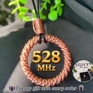 Könnte beinhalten: Ein kreisförmiger Anhänger aus gedrehtem Kupferdraht, der an einer schwarzen Schnur hängt. In der Mitte des Rings befindet sich der goldene Text 528 MHz. Ein kleiner runder Einsatz zeigt eine Geschenkbox mit einem Ring und dem Text Gift for you 333MHz.