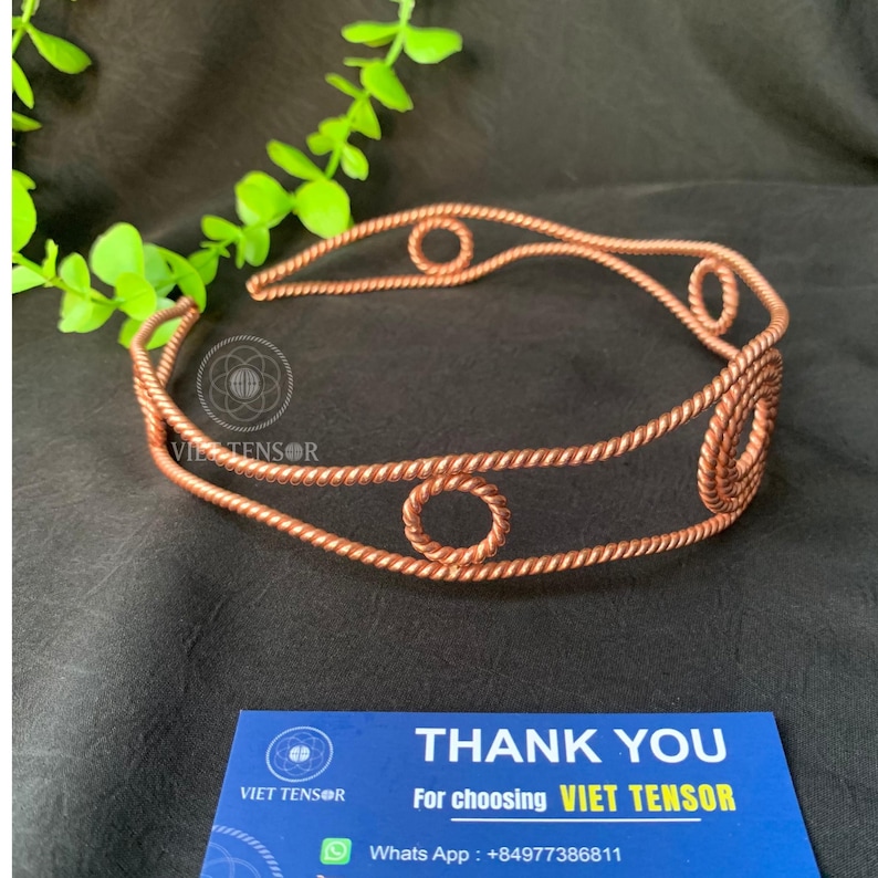 May include: A copper-colored wire headband featuring a twisted rope texture and circular accents. The headband is displayed alongside a blue business card that reads THANK YOU For choosing VIET TENSOR and includes a WhatsApp number +84977386811.
