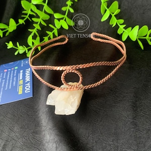 May include: A copper-colored, open-ended bracelet with a twisted rope design. The bracelet has a circular design in the center and rests on a white crystal. The background is black with green foliage.