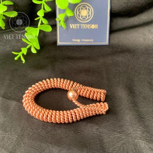 May include: A copper-colored bracelet with a coiled design and a spherical accent. The bracelet is made of tightly wound wire and features a smooth, round bead. The background includes a card with the text "VIET TENSOR" and "Energy - Frequency."