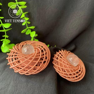 May include: Two copper-coloured wire art pieces, each with a central, clear, rounded stone. The wire is woven in a spiral pattern. The pieces are on a dark background. The text "VIET TENSOR" is in the upper left corner.