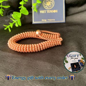 May include: A copper-colored coiled wire bracelet featuring a single spherical bead at the closure. The bracelet is displayed near a dark blue card labeled VIET TENSOR Energy-Frequency and a circular graphic with the text GIFT for you 333MHz.