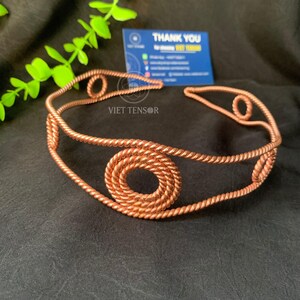 May include: A copper-colored metal headband featuring a twisted wire design. The headband is constructed from two parallel twisted wires connected by three circular motifs, each formed by a continuous coil of the same twisted copper-colored metal wire.