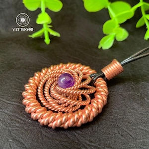Puede incluir: Colgante de cobre con diseño en espiral, con una gema púrpura central. El colgante cuelga de un cordón negro. El logotipo "VIET TENSOR" es visible. El colgante está sobre un fondo oscuro con follaje verde.