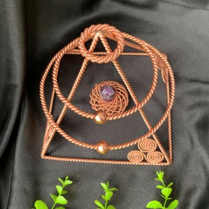 Pode incluir: Uma escultura piramidal de fio de cobre com textura de corda trançada. A estrutura inclui uma bobina circular central que segura uma pequena esfera de ametista roxa, duas esferas de cobre lisas e três espirais na base.