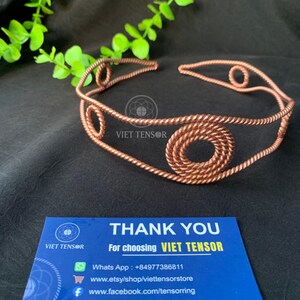 May include: A copper wire headband featuring a twisted rope texture and circular spiral accents. The headband is accompanied by a blue business card that reads THANK YOU For choosing VIET TENSOR, including contact details and social media links.