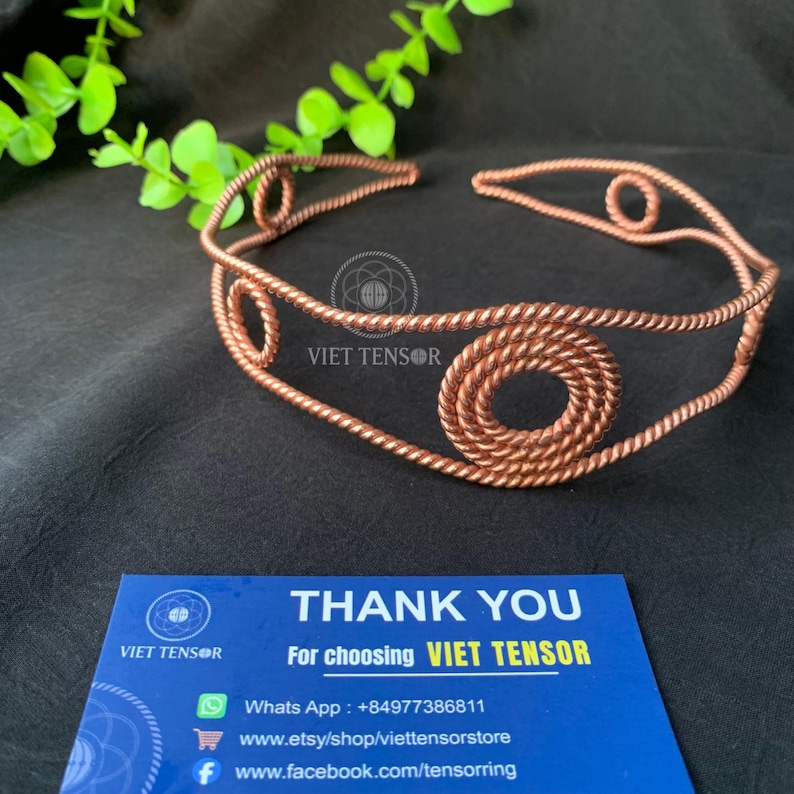 May include: A copper wire headband featuring a twisted rope texture and circular spiral accents. The headband is accompanied by a blue business card that reads THANK YOU For choosing VIET TENSOR, including contact details and social media links.