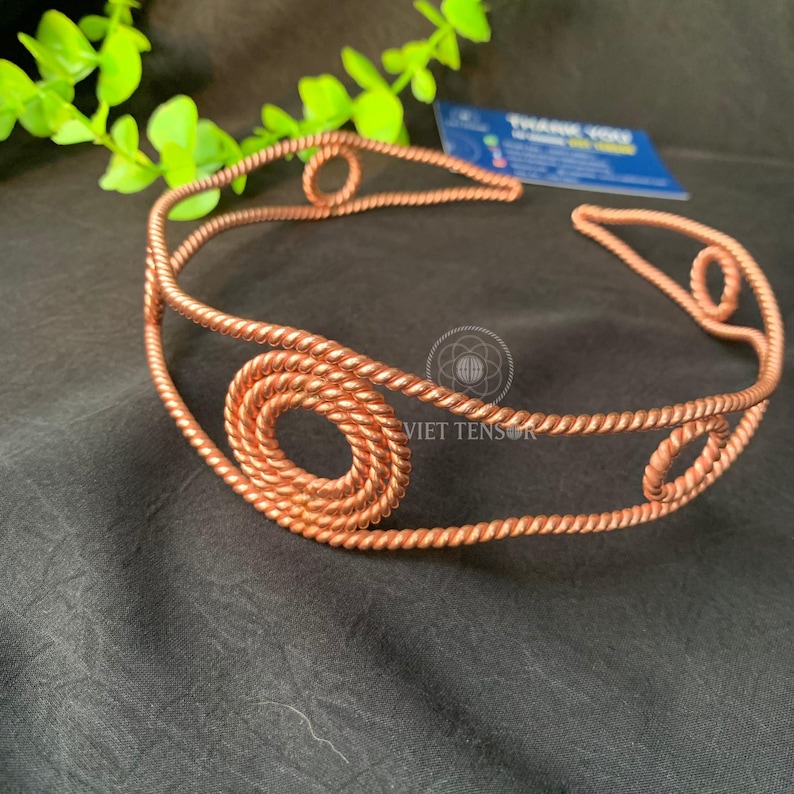 May include: A copper-colored metal headband constructed from twisted wire. The design features a series of circular, coiled wire motifs integrated into the band, which consists of two parallel twisted wire strands. The metal has a warm, metallic finish.