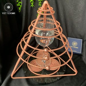 Könnte beinhalten: Eine kupferfarbene geometrische Skulptur mit einem Glas, das Wasser enthält. Die Skulptur hat eine dreieckige Basis und ein spiralförmiges Design um das Glas. Der Text "VIET TENSOR" ist oben links und auf einer blauen Box zu sehen.