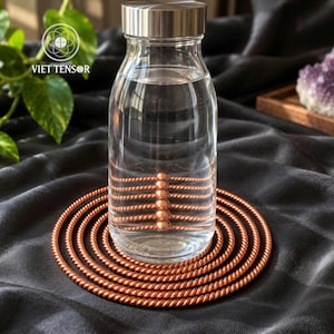 May include: A clear glass water bottle with a silver-toned metal cap sits atop a circular, copper-colored coiled wire coaster. The coaster features a repeating twisted texture. The logo VIET TENSOR is visible in the upper left corner.