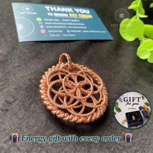 May include: A circular copper pendant featuring an intricate, repeating geometric pattern of interconnected loops and twisted wire strands. The design forms a symmetrical, flower-like structure with a small bail at the top for suspension.