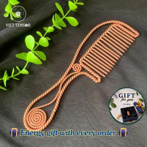 Könnte beinhalten: Ein kupferfarbener Kamm aus Draht mit einem gedrehten Metalldesign und einer kreisförmigen Spirale am Griffende. Der Kamm verfügt über eine Reihe paralleler Zinken und ein dekoratives Knotendetail an der Verbindung zwischen Griff und Kammkopf.
