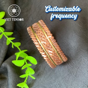 Può includere: Braccialetti in rame con un design strutturato e dettagli color oro. I braccialetti sono impilati e mostrati su una superficie grigio scuro. Il testo "Customizable frequency" è visibile.