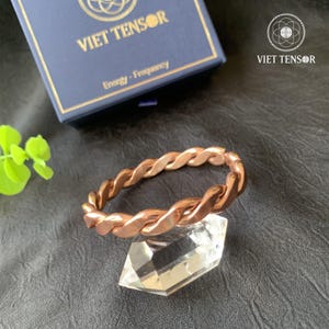 May include: A copper-coloured twisted metal bracelet rests on a clear crystal, with a dark blue box in the background. The box has the text "VIET TENSOR" and "Energy - Frequency". The bracelet is a continuous loop.