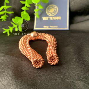 May include: A copper-colored, horseshoe-shaped object with a central sphere. The object is made of tightly woven strands, with the text "VIET TENSOR" visible in the background. The item is displayed on a dark surface.