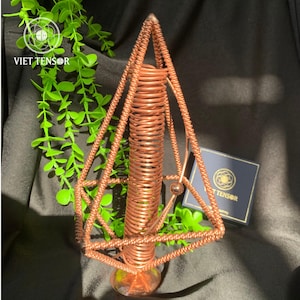 Puede incluir: Un tensor de color cobre con un diseño geométrico. El tensor presenta una bobina central y un marco en forma de diamante. La base tiene una forma clara y redondeada. El producto se muestra con vegetación y una caja con el texto "VIET TENSOR".