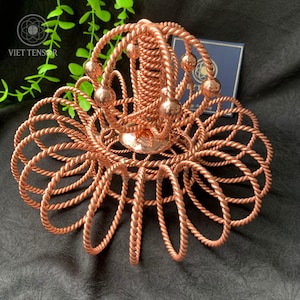 Può includere: Un oggetto decorativo fatto a mano, color rame, con un design floreale. Il pezzo presenta una colonna centrale a spirale, circondata da fili ad anello simili a corde. Accenti sferici sono uniformemente distanziati sulla parte superiore. Il logo "VIET TENSOR" è visibile.