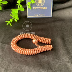May include: A copper-colored bracelet with a woven design and a small spherical bead. The bracelet is displayed on a dark surface, with a card in the background that reads "VIET TENSOR" and "Energy - Frequency".