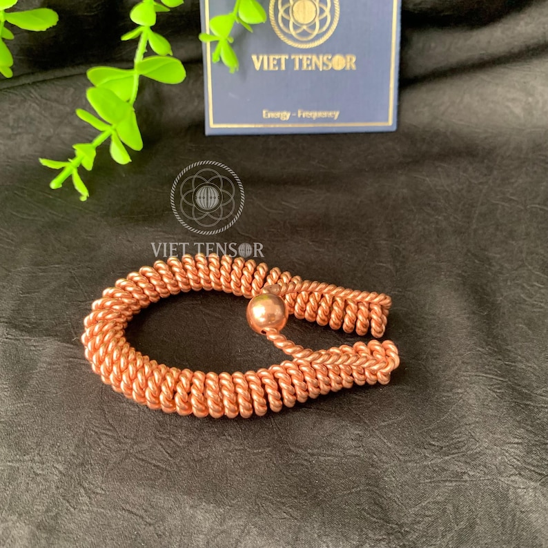 May include: A copper-colored bracelet with a woven design and a small spherical bead. The bracelet is displayed on a dark surface, with a card in the background that reads "VIET TENSOR" and "Energy - Frequency".