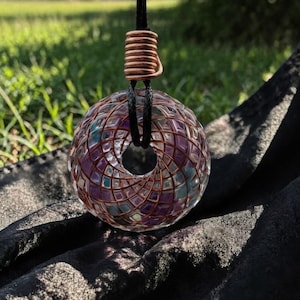 Può includere: Un ciondolo sferico trasparente con un motivo geometrico di quadrati viola, blu e trasparenti, racchiuso in filo di rame. Pende da un cordino nero con un accento a spirale di rame. Lo sfondo è erba verde sfocata e tessuto scuro.