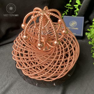 Peut inclure: Une œuvre d'art artisanale de couleur cuivrée avec un motif en spirale. La pièce présente une structure tissée en forme de panier avec un cristal clair central et des accents sphériques. La base est sombre et texturée, avec le texte "VIET TENSOR".