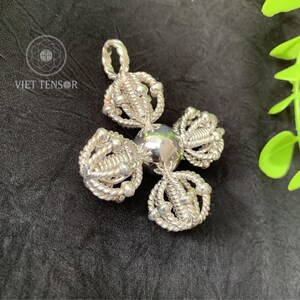 May include: A silver pendant with a unique, four-petal design. Each petal is crafted from intricately woven silver wire, adorned with small, round silver beads. The central element is a polished silver sphere. The pendant is displayed on a black surface.