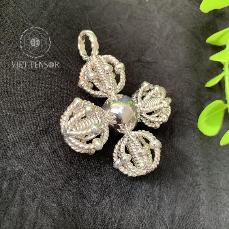 May include: A silver pendant with a unique, four-petal design. Each petal is crafted from intricately woven silver wire, adorned with small, round silver beads. The central element is a polished silver sphere. The pendant is displayed on a black surface.
