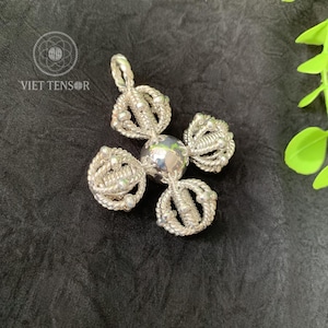 May include: A silver pendant with a central sphere and four ornate, looped arms. The pendant is made of woven silver strands and small silver beads. The text "VIET TENSOR" is visible in the upper left corner.