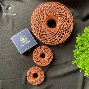 Peut inclure: Vue aérienne de trois objets en spirale de couleur cuivrée, avec une boîte portant l'inscription "VIET TENSOR" en lettres dorées. Les objets sont disposés sur une surface sombre et texturée, avec un petit bouquet de feuillage vert.