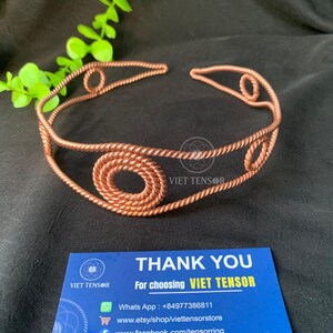 May include: A copper-colored metal headband crafted from twisted wire, featuring a series of circular coiled accents along the band. The piece is displayed alongside a blue card with the text THANK YOU For choosing VIET TENSOR.