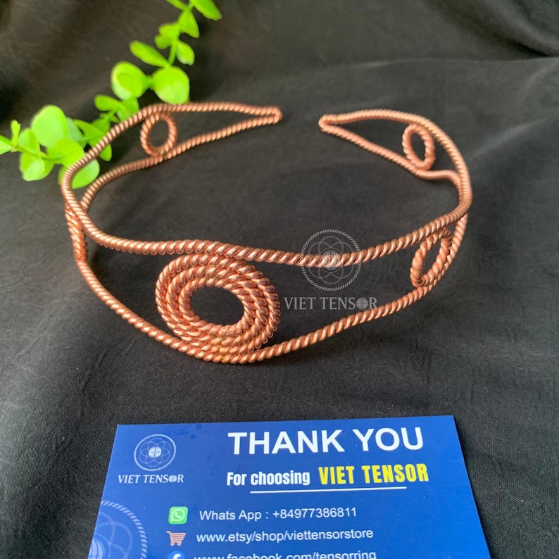 May include: A copper-colored metal headband crafted from twisted wire, featuring a series of circular coiled accents along the band. The piece is displayed alongside a blue card with the text THANK YOU For choosing VIET TENSOR.