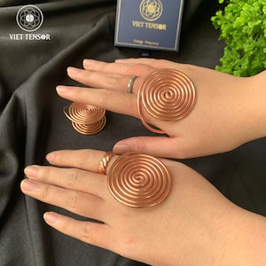 Peut inclure: Anneaux en spirale en cuivre et un petit pendentif en spirale sur fond sombre. Les anneaux sont de tailles différentes, dont un grand anneau en spirale sur un doigt. Une boîte avec le texte "VIET TENSOR" est visible.