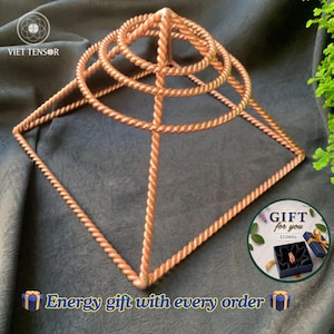 Op de afbeelding: Een koperkleurige draadpiramidestructuur met een vierkante basis en drie concentrische cirkelvormige ringen aan de bovenkant, allemaal gemaakt van gedraaid metaaldraad. Het object bevat een kleine ronde grafische inzet met een ring in een doosje.