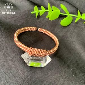 Op de afbeelding: Een koperkleurige armband met een helder kristallen accent. De armband heeft een gedraaid touwontwerp en is open. Het kristal is facetgeslepen en heeft een geometrische vorm. De armband wordt gepresenteerd op een donkere ondergrond.