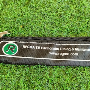 Op de afbeelding: Zwarte etui met ritssluiting en witte tekst "RPGMA TM Harmonium Tuning & Maintenance KIT" en "www.rpgma.com". De etui heeft een groen-wit logo. De achtergrond is groen gras.