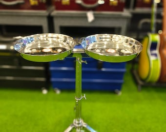 May include: A shiny, chrome-plated percussion instrument, likely a set of tabla drums, with two bowl-shaped drums mounted on a tripod stand. The drums reflect the surrounding environment. The stand has adjustable height and a three-legged base.