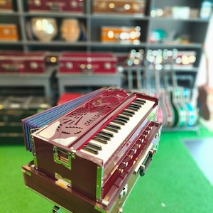May include: A maroon and silver harmonium with black and white keys, displayed on a silver stand. The instrument has the word "HARMONIUM" engraved on the front. The background shows shelves with other musical instruments.