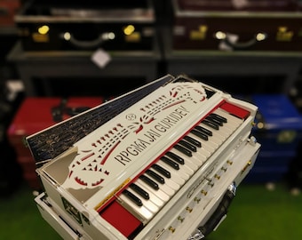 May include: A white harmonium with black and white keys, red accents, and the text "RPGMA JAI GURUDEV". The instrument has a handle and is displayed in front of a shelf with other similar instruments.