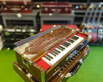 Könnte beinhalten: Ein hölzernes Harmonium mit einer Tastatur, roten Akzenten und kunstvollen Schnitzereien. Das Instrument steht auf einem Ständer, mit einem Griff und silbernen Zierleisten. Weitere Musikinstrumente sind im Hintergrund zu sehen.