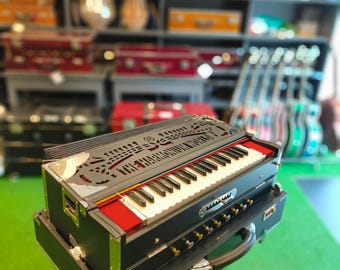 Pode incluir: Um harmônio preto e prateado com teclas brancas e detalhes vermelhos. O instrumento musical está em um suporte, com uma alça e vários botões. O fundo mostra prateleiras cheias de outros instrumentos musicais.