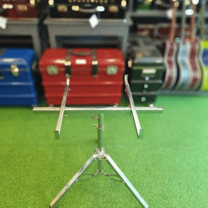 May include: A silver-colored, adjustable music stand with a tripod base. The stand has a horizontal bar with two angled supports. The background features a variety of cases and guitars, suggesting a music store setting.