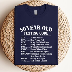 May include: Navy blue t-shirt with white text: "80 YEAR OLD TEXTING CODE" and a list of acronyms with their meanings. The shirt is folded on a woven, circular surface.