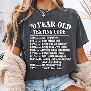 May include: Dark gray t-shirt with the text "70 YEAR OLD TEXTING CODE" in white. The shirt lists common texting abbreviations with their meanings, such as "ATD - At The Doctor" and "BFF - Best Friend Fell".