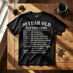 May include: Black t-shirt with white text that reads "50 YEAR OLD TEXTING CODE" and a list of acronyms with their meanings. Sunglasses and a cup of coffee are on a wooden table.