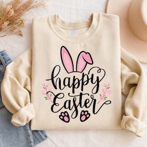 May include: A cream-colored sweatshirt featuring a "Happy Easter" design. The design includes pink bunny ears, floral accents, and bunny paw prints. The text is in a stylish script font, perfect for Easter celebrations.