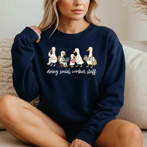 May include: Navy blue sweatshirt with a white graphic of ducks in various social worker roles. The text "doing social worker stuff" is below the ducks in a handwritten style font.